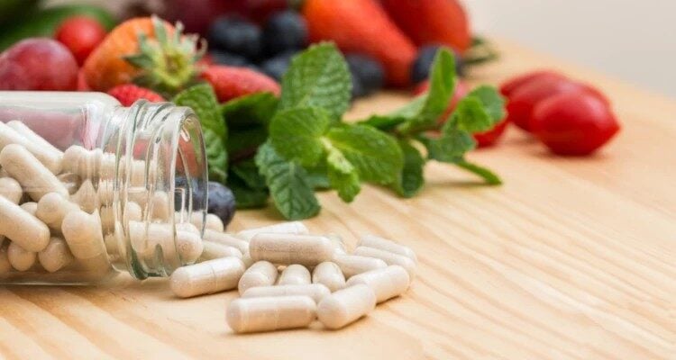 Staklena bočica iz koje ispadaju kapsule s vitaminima i mineralima. U pozadini se nalazi voće i povrće