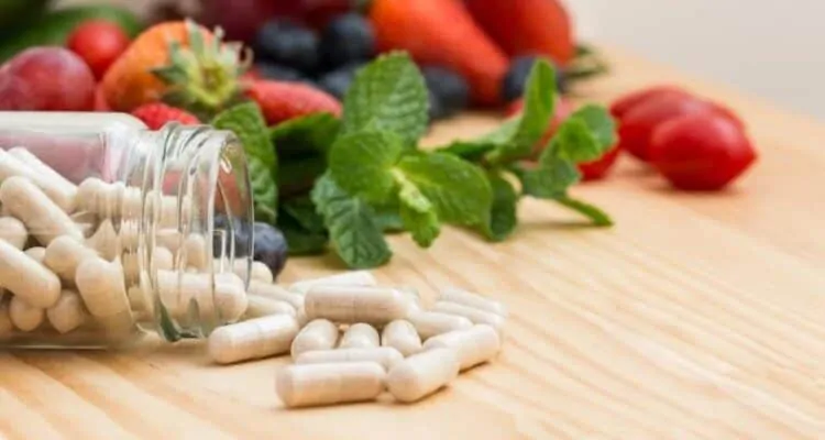 Staklena bočica iz koje ispadaju kapsule s vitaminima i mineralima. U pozadini se nalazi voće i povrće